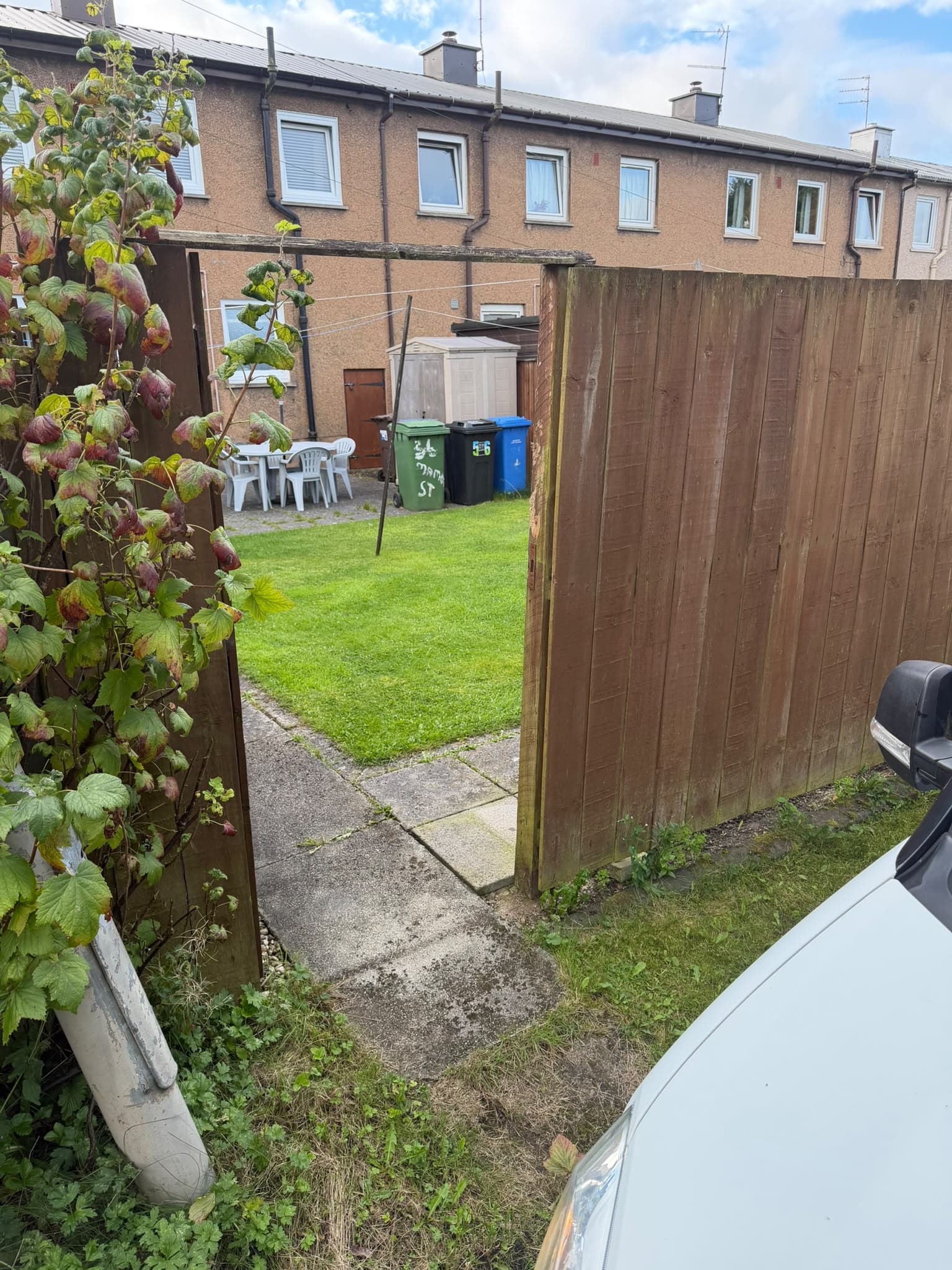 Gap in the failed boundary fence looking straight into the rear garden, drying green and bins from the service lane