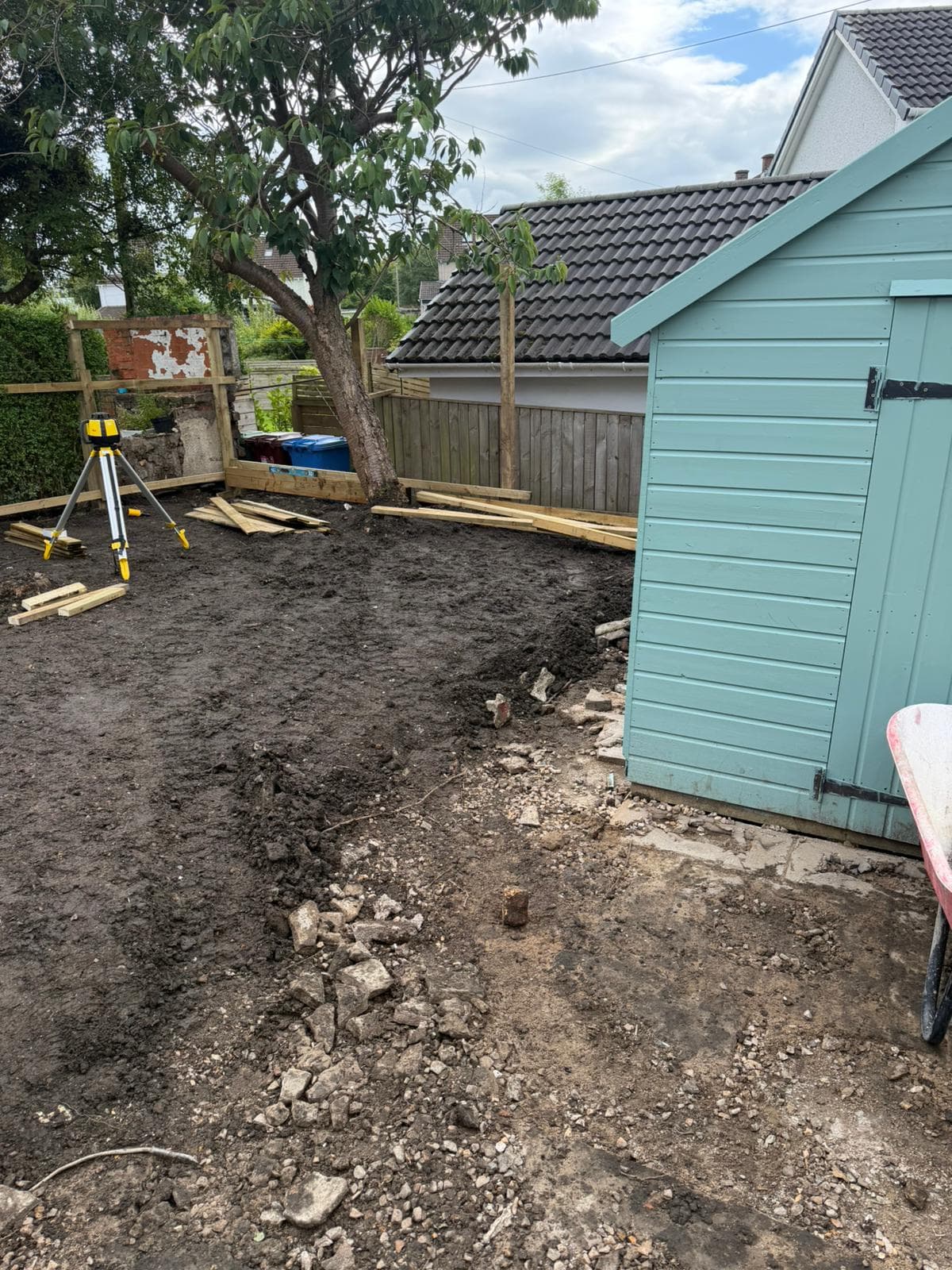 Rear garden mid-excavation with a laser level set up, soil stripped back and rubble exposed beside a garden shed
